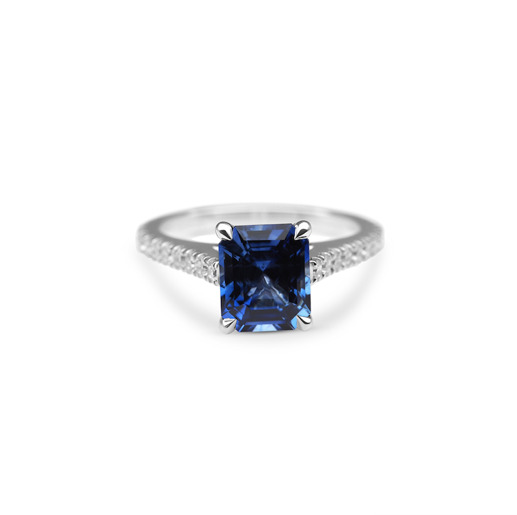 White gold ring with an emerald-cut blue sapphire gemstone and diamond-encrusted band on a white background.