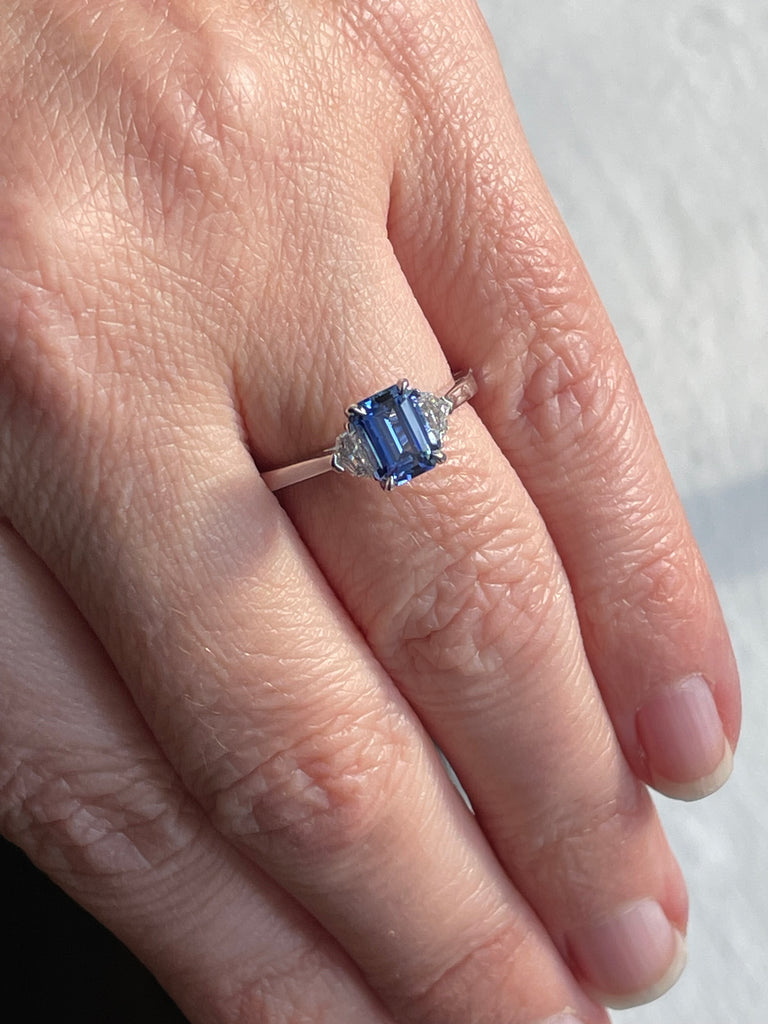 A hand wearing a ring with a large blue emerald cut sapphire gemstone framed by smaller white diamonds on either side