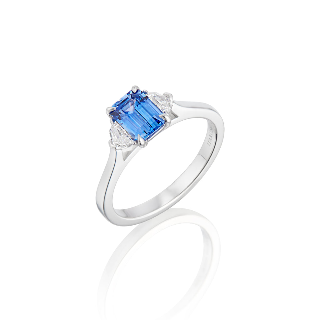 White gold ring with an emerald-cut blue sapphire flanked by two smaller diamonds on a reflective surface.