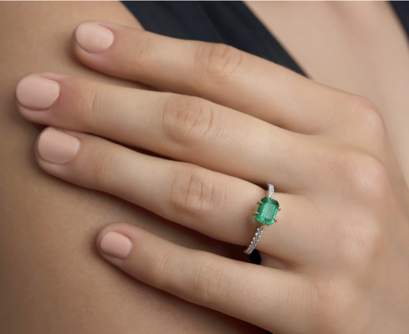 Hand with manicured nails wearing a ring with a green emerald centre and diamond accents.