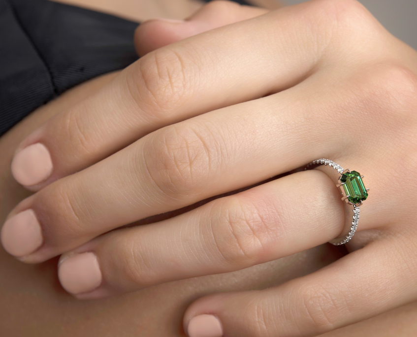 Hand wearing a ring with a green gemstone and diamond accents.