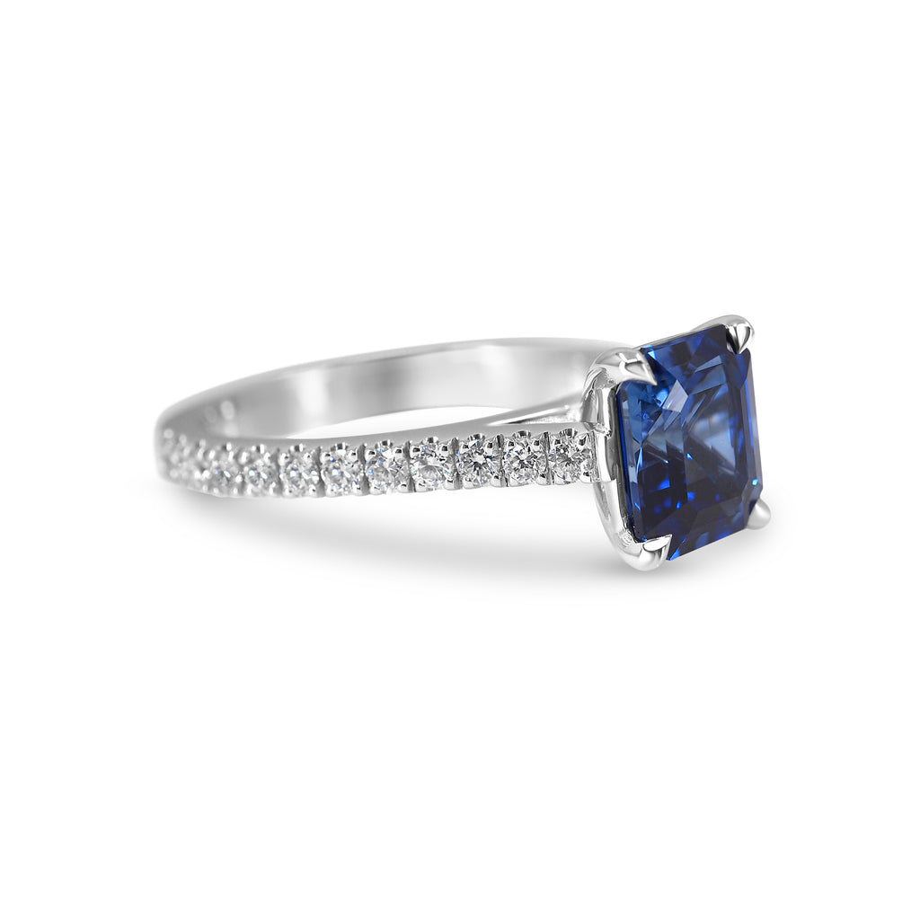 White gold ring with an emerald-cut blue sapphire and small diamonds on the band, isolated on a white background.