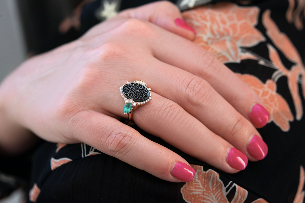 A hand with pink nail polish wearing a ring with a black filigree pattern and emerald and diamond gemstones