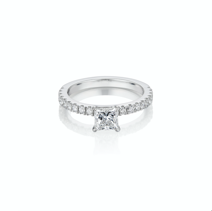 A pristine princess cut diamond engagement ring with pavé set round diamonds on the band on a white background.