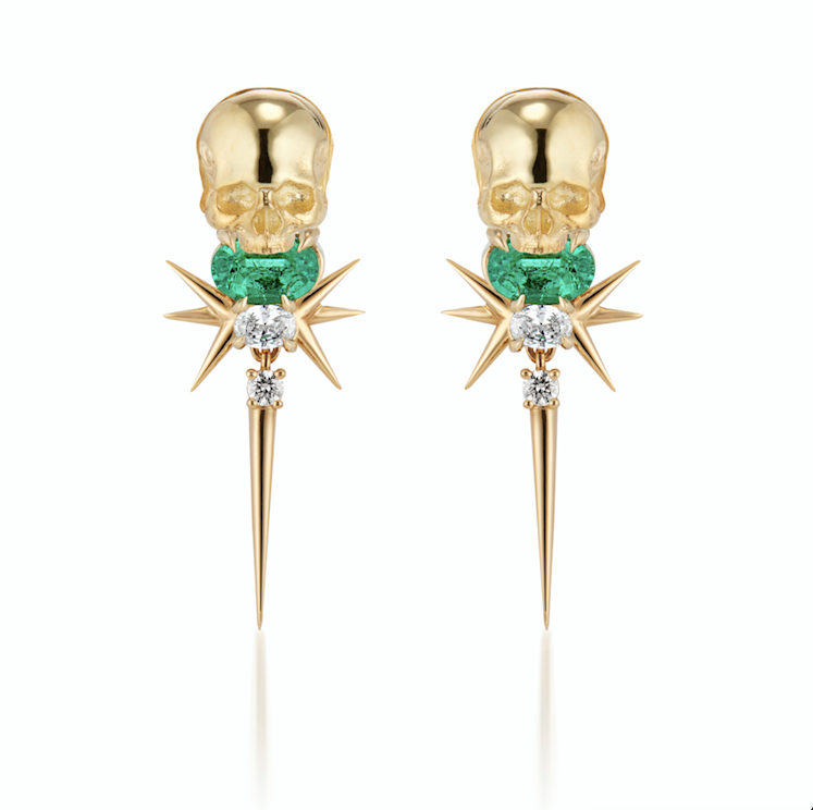 A pair of golden skull earrings with green emerald gems and diamond accents on starburst designs.