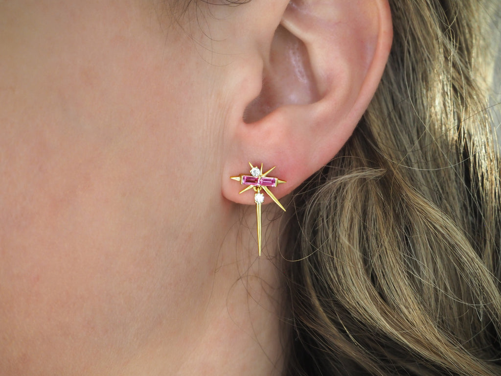 Close-up of an ear wearing a decorative gold cross earring with a pink gem.