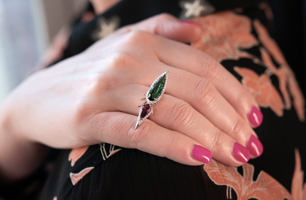 Closeup on a woman’s hand wearing a fine jewelry piece, the Tourmaline Diamond Ring on her ring finger. The pink tourmaline gem extends halfway over the pinky and the green tourmaline gem extends halfway over the middle finger. The tourmaline gems join together slightly off center for an asymmetrical look. The ring sits in the middle of the first knuckle.