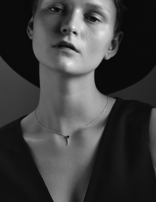 A black-and-white image of a person wearing a hat and a necklace with a blue pendant.